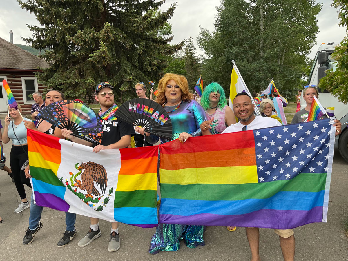 A group of smiling people dressed in colorful clothing, makeup and wigs walk for Avon's Mountain Pride parade and hold up two tapestry flags.