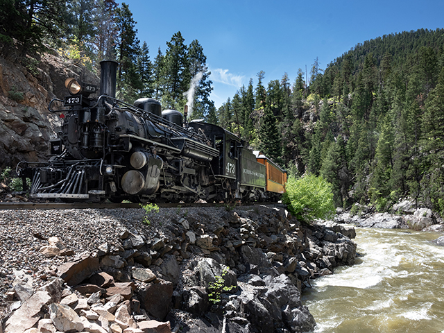 The Durango Train chugs along its track beside a rushing river. Along the sides of the tracks are small cliffs of sharp rocks. In the background is a large hill with pines.