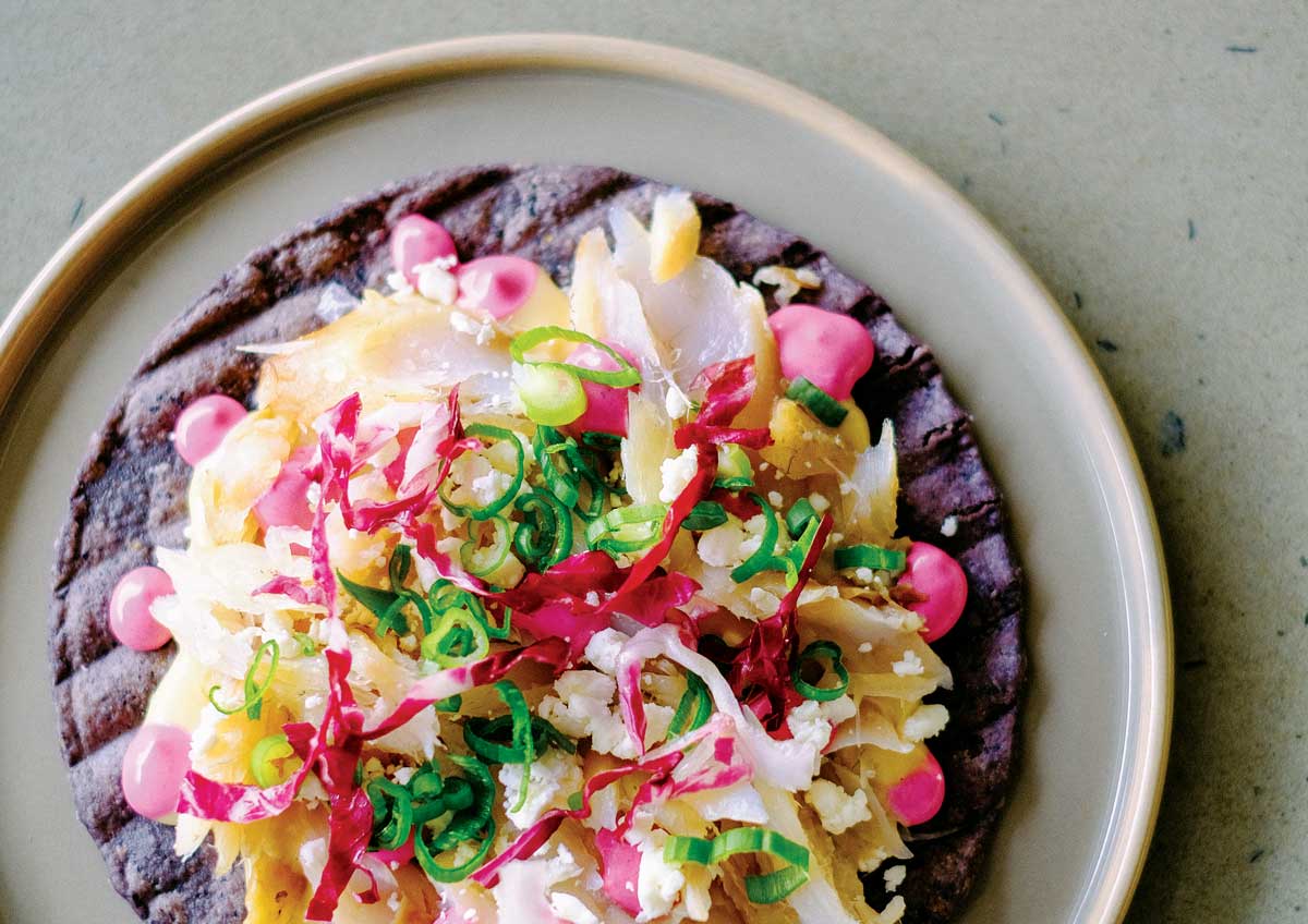 A blue-corn tortilla sits on a white, ceramic plate on a table in Colorado and is topped with red beans, squash blossoms, green onions and other vibrantly colored ingredients.