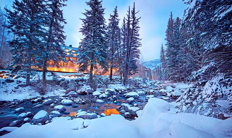 A river filled with snow-covered rocks runs by the warmly lit hotel