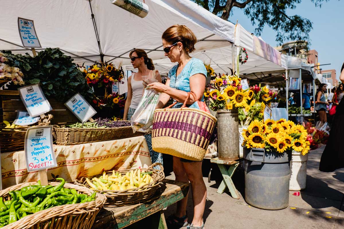 Two women holding baskets peruse pepper produce in a farmers' market booth