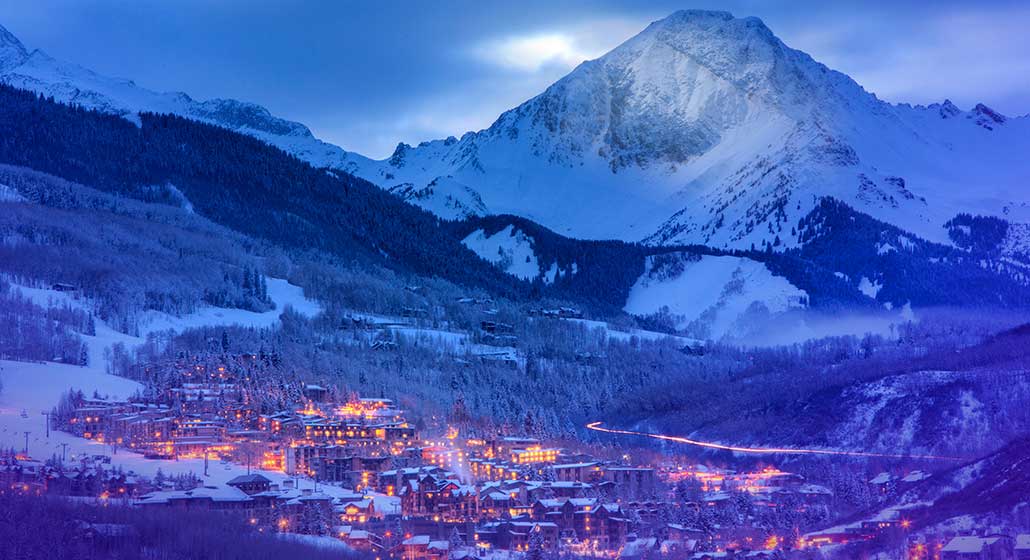 An aerial view of Snowmass Village nestled at the base of a Rocky Mountain pass. The whole town and mountains are covered in snow.