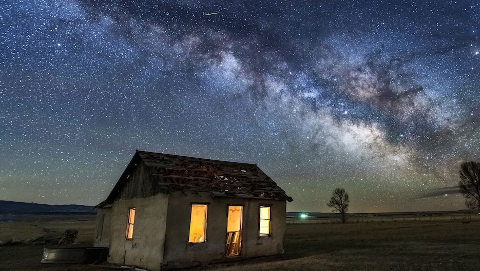 A bright display of the Milky Way and stars above an old cabin in a grassy field