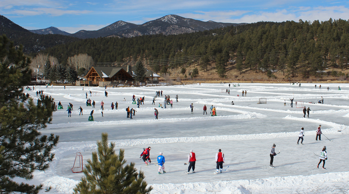 People in hockey and skating gear enjoy themselves on the white rink of the Evergreen Lake in the winter. In the background are mountains and groves of pine trees, underneath mostly blue skies.