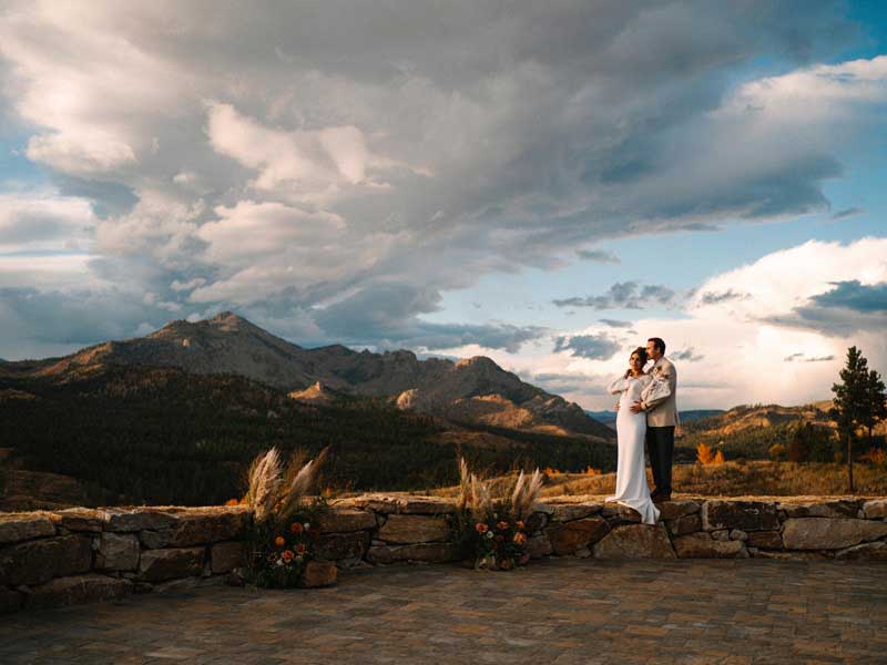 A just-married couple in white dress and suit gaze at fluffy clouds moving over a mountain ridge