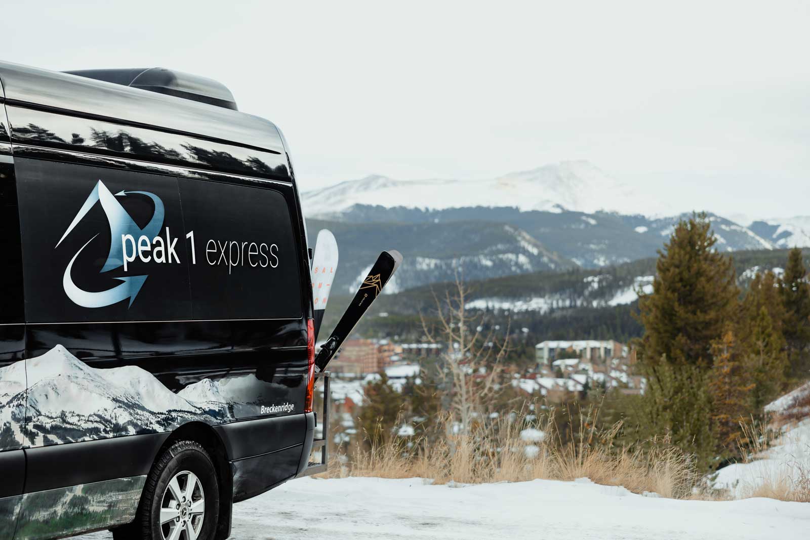 A black sprinter van with skis on the back; logo reads Peak 1 Express