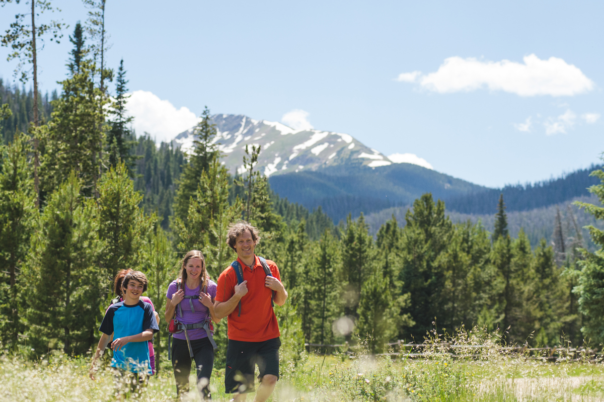 A family of four walks and smiles next to each other on a hike in Winter Park, Colorado. All around them are fields of grass and wildflowers, and in the background, the viewer can see out-of-focus towering mountain peaks.