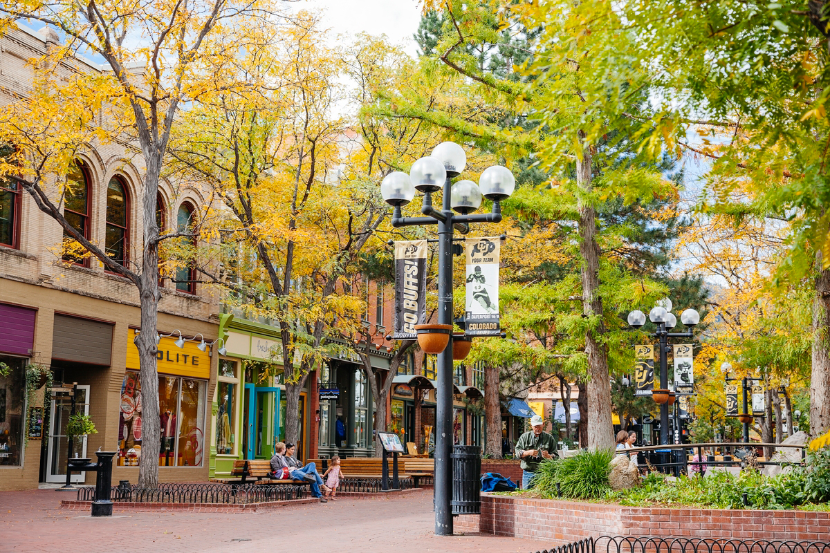 A photo of Pearl Street in Boulder, Colorado, during the fall. There are trees whose leaves are turning yellow beside brick storefronts in front of the cobblestone walking path, and in the middle of the photo is a light post with flyers that say "GO BUFFS!" to celebrate Colorado University.