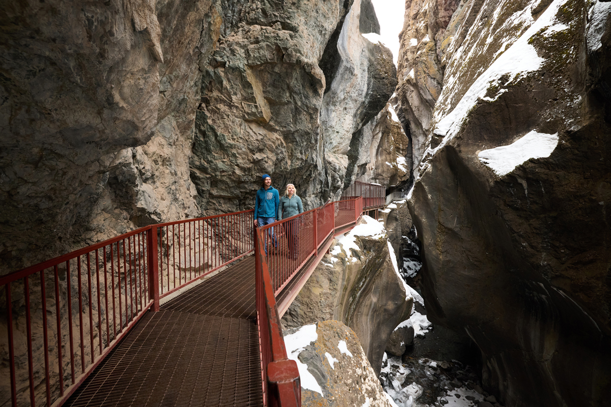 Two people walk along a bridge past snow covered rock walls at Box Cañon Falls in winter in Ouray, Colorado