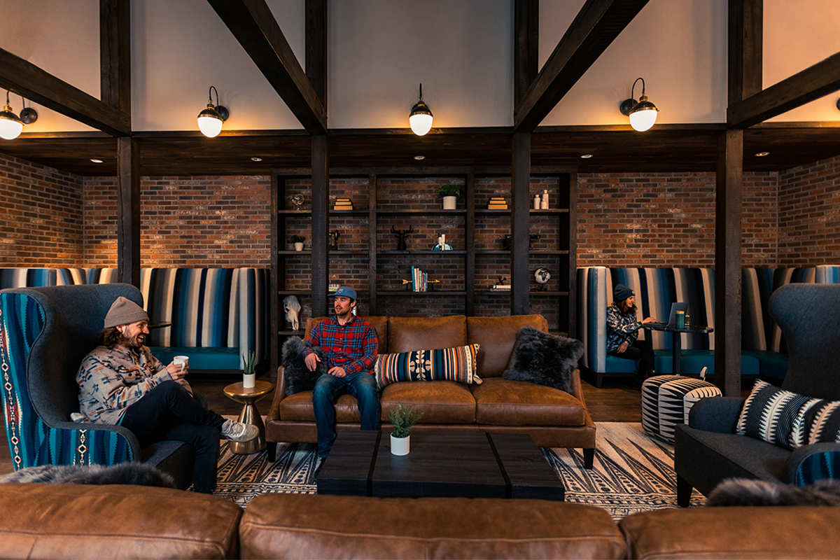 Three people share the downstairs community space at The Bivvi Telluride in Colorado. One works on their computer in a blue, white and black-striped nook the other two sit and chat around a dark coffee table.