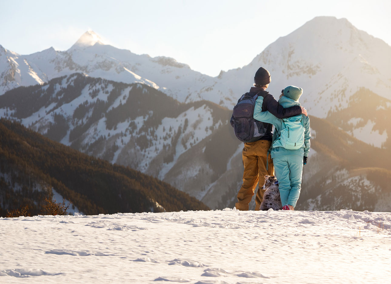 Two people with a pup at their feet stand on in a snowy field and look at snowcovered peaks