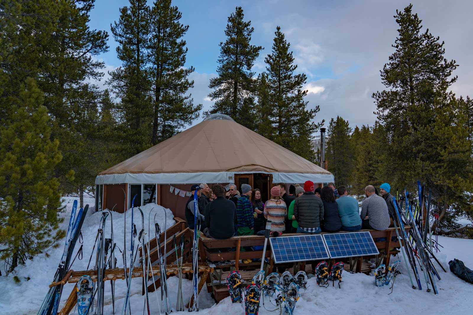 A group of people socialize and get warm at a yurt while surrounded by cross-country skis pointed at the sky