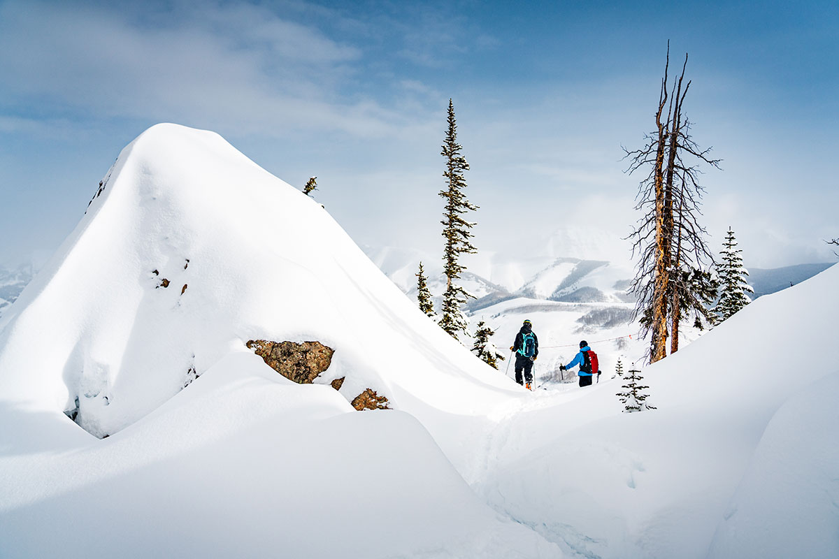 Snow-covered rock with two skiers behind it and mountains in the distance
