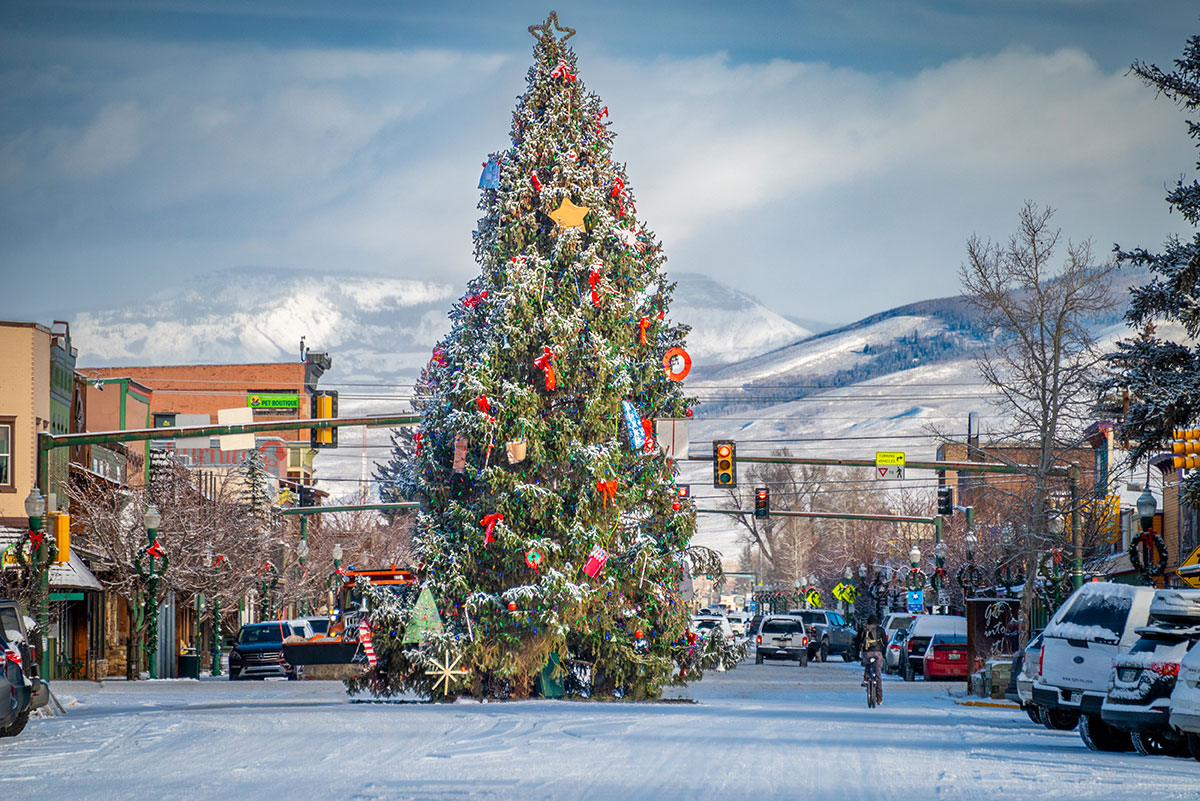 A very tall Christmas tree decorated with ribbons in the middle of a snowy, small-town street