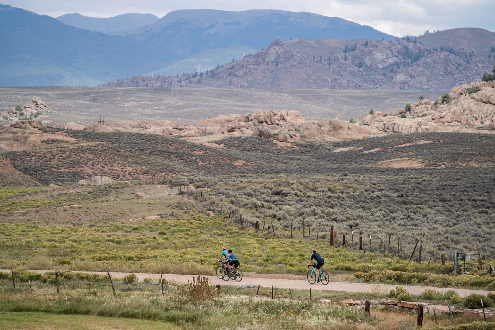 Three gravel bikers ride a dirt road past red-rock formations that sit behind a fence