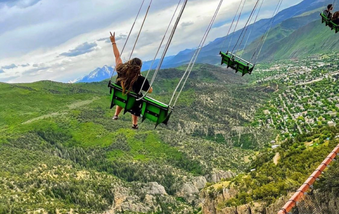 A picture shot from behind  of a person in a swing making a peace sign as they soar over a lush canyon on the Glenwood Caverns Flyer at Glenwood Caverns Adventure Park in Glenwood Springs, Colorado.
