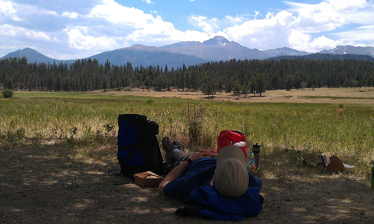 Man laying on the ground in the shade wearing a tan hat with mountain peaks and forest trees ahead of him