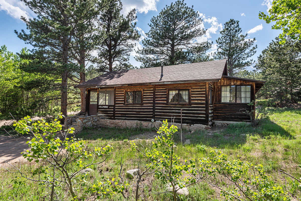 A log cabin at Meeker Park Lodge sits on a lovely wooded lot with tall pine trees and a cloud-streaked blue sky behind it.