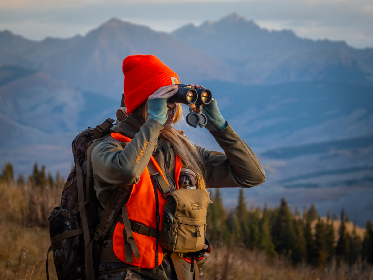 A hunter in a bright orange cap and vest looks through binoculars in the Gunnison Valley of Colorado.