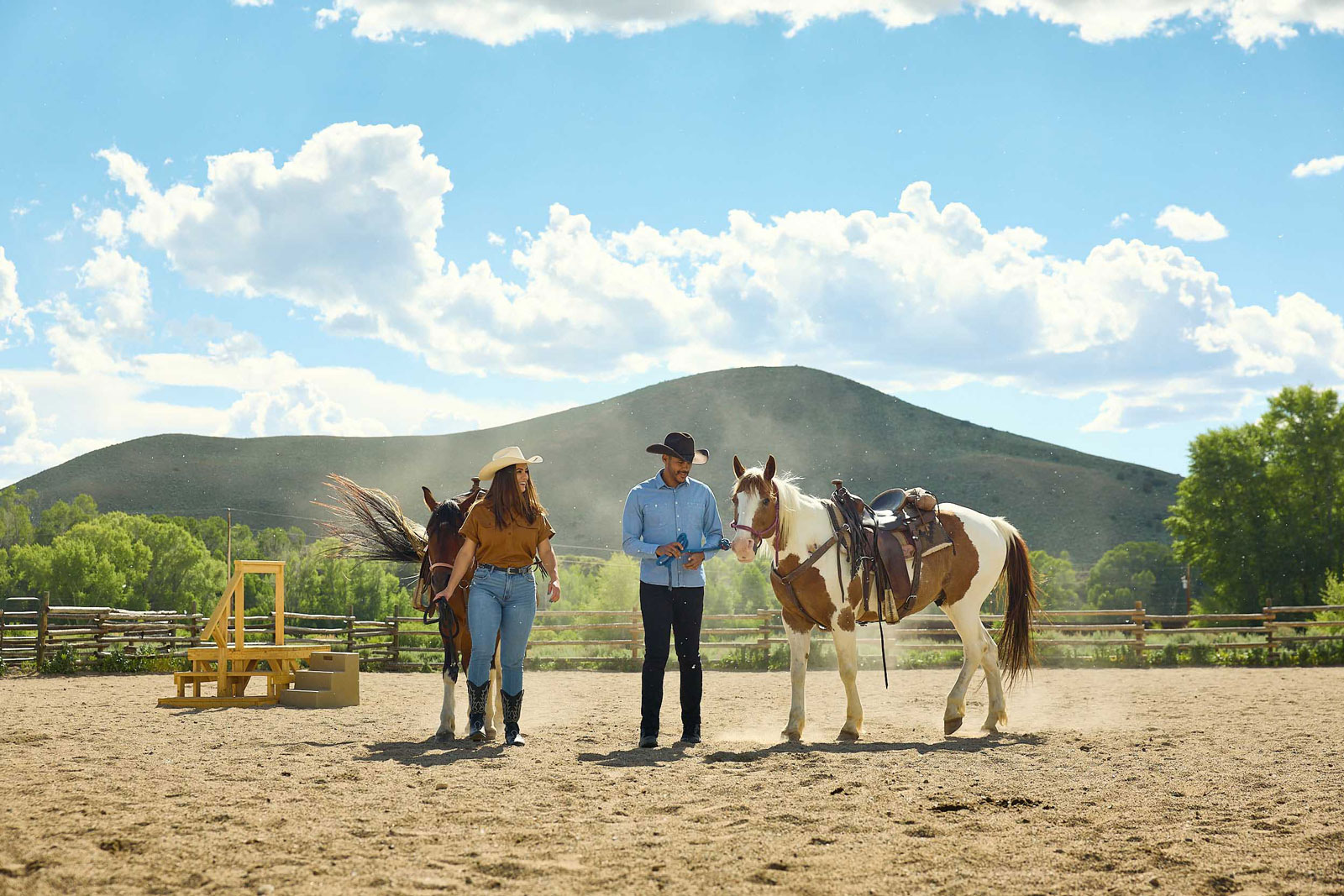Two people in cowboy boots and hats lead saddled horses through a corral