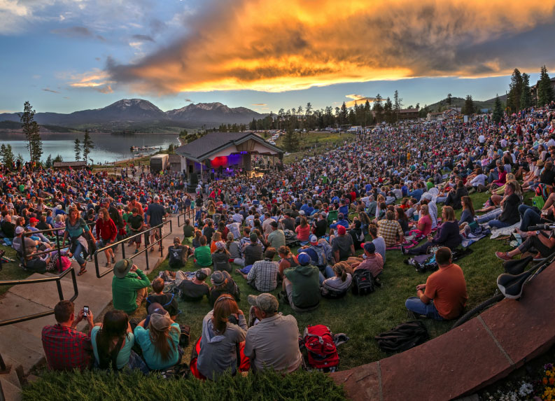 A crowd sits on the lawn before lakeside Dillon Amphitheater as the sun sets behind the mountains