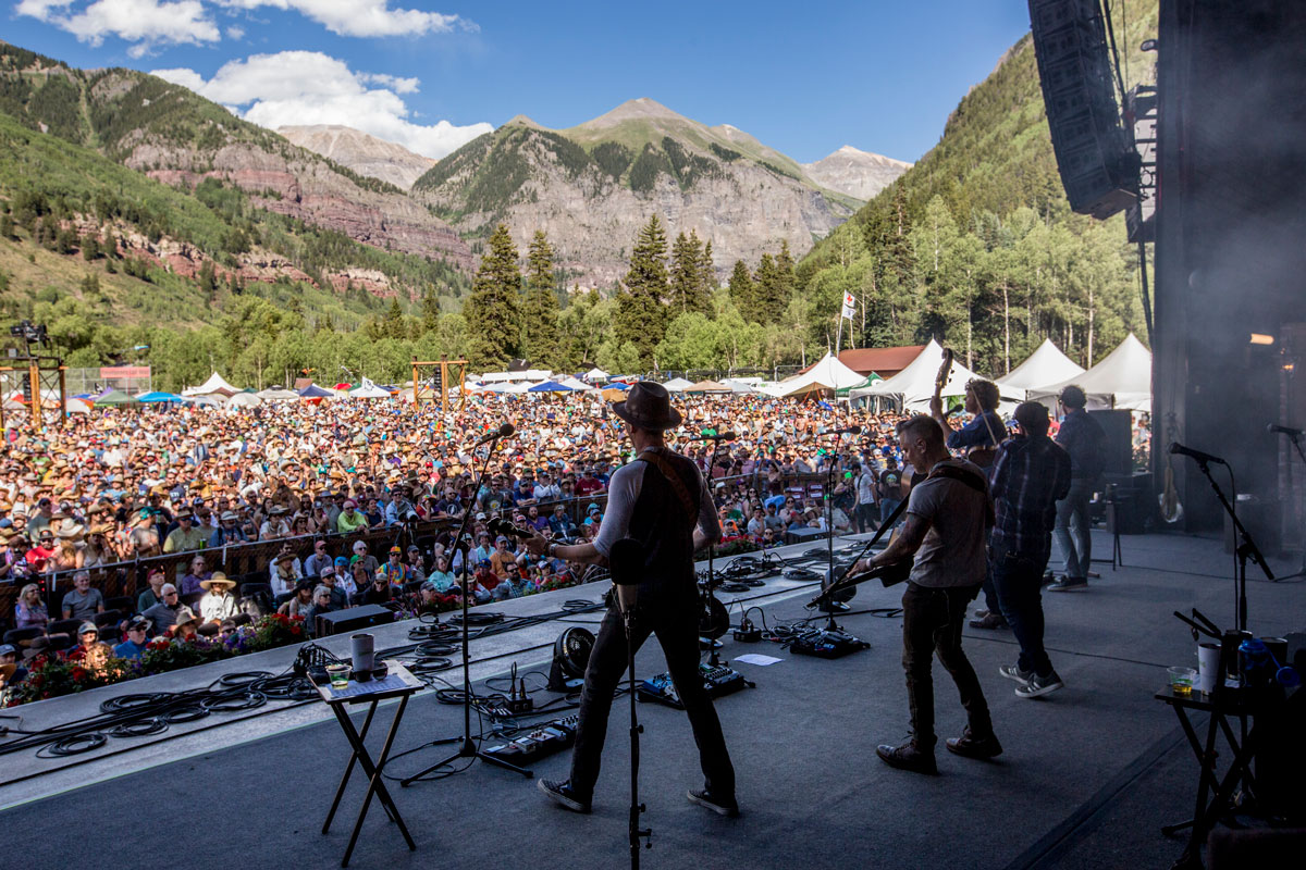 A band plays on stage and looks out to a crowd and mountain scene