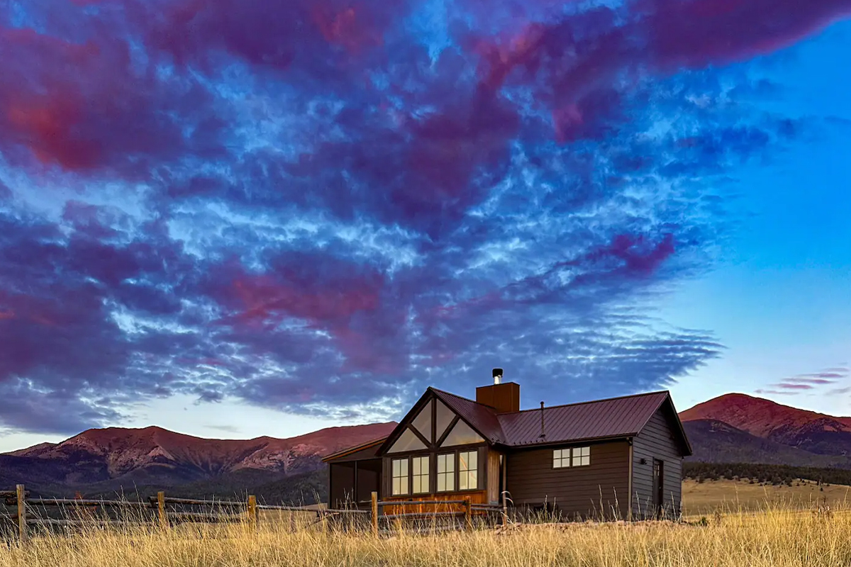 The cabin at Three Peaks Ranch outside Westcliffe, Colorado, stands out against the mountains, blue sky and purple-tinged clouds in the background.