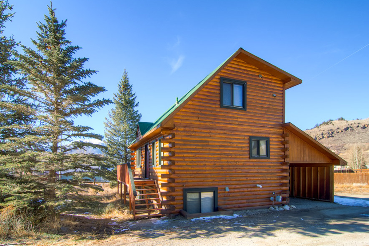 A two-story log cabin with a covered garage and a deck is set near tall pines and rolling foothills in Dillon, Colorado.