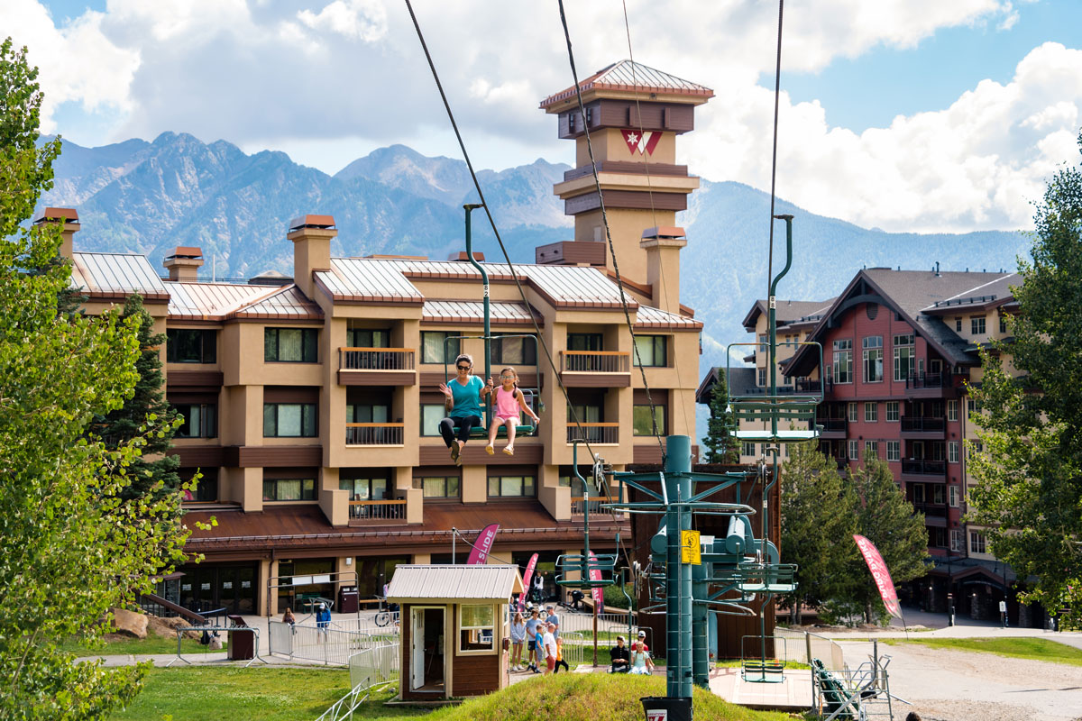 Mother and daughter riding a chairlift, ascending toward the sky with Purgatory Resort in the background.