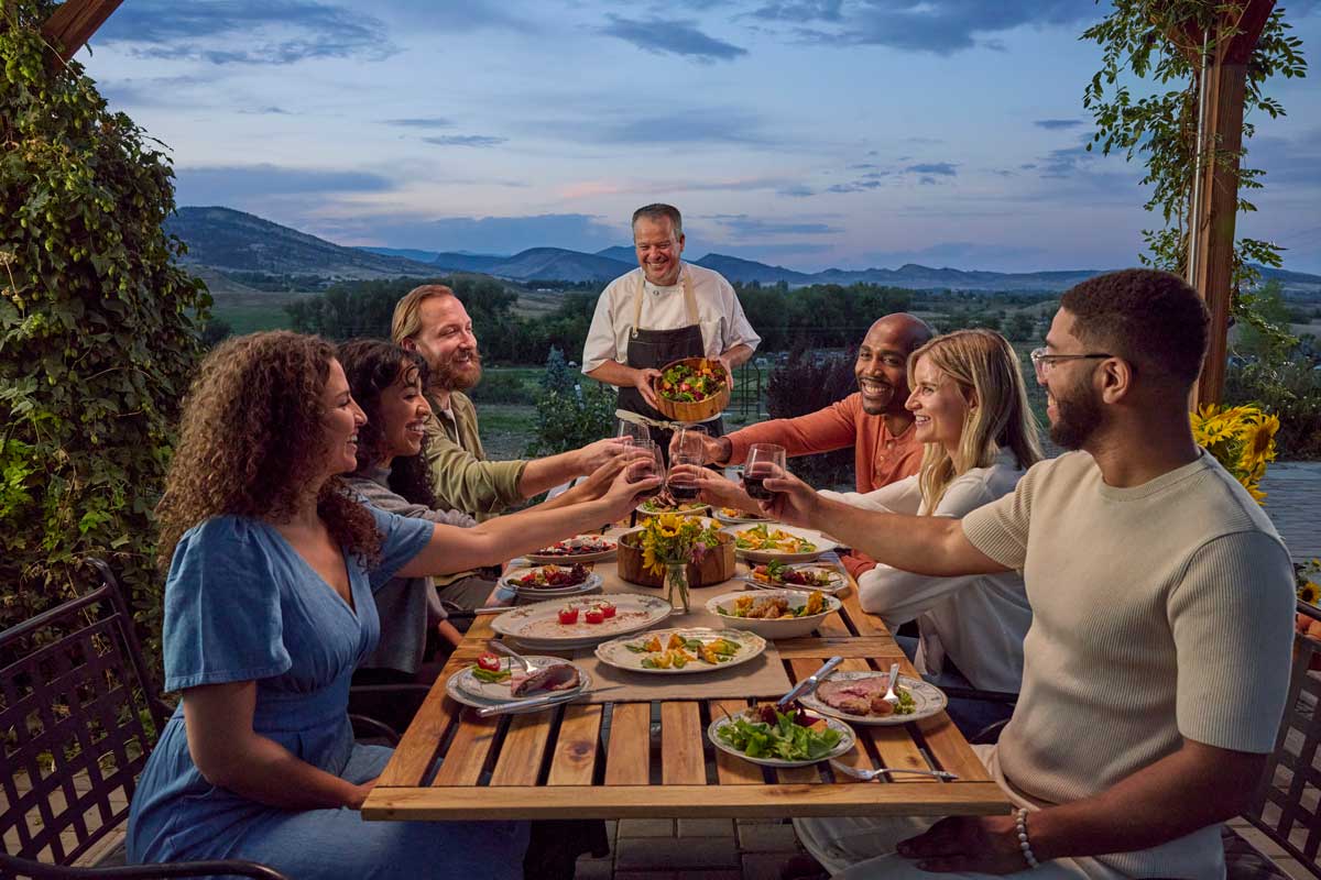 A group of people at a table in the field of a farm share a toast to the chef