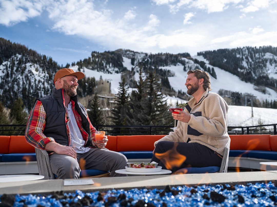 Two men drink glasses of wine on a patio with a Coloado ski resort in the distance