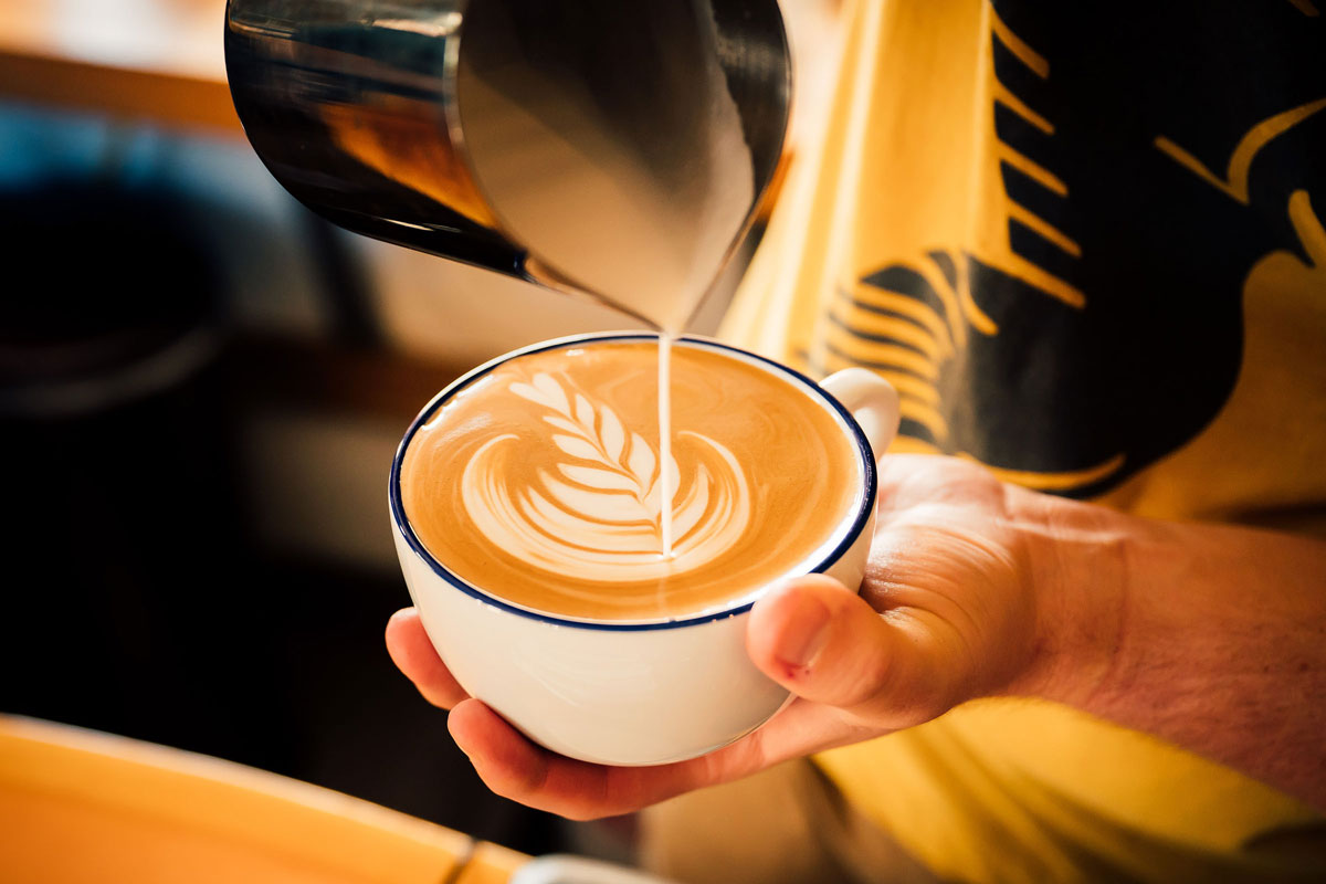 Close up of a barista's hand holding a latte, creating a design on top of the drink.