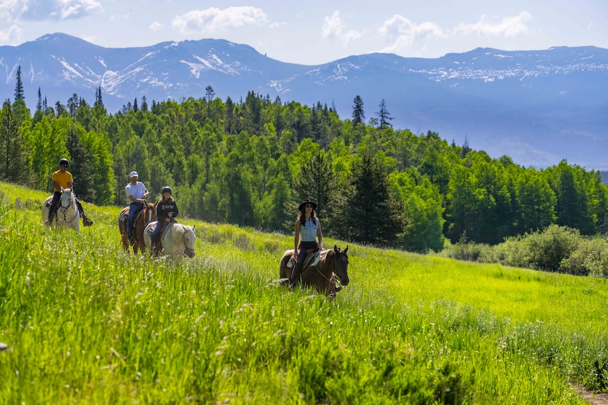 Adults riding horseback through a green meadow with mountains in the background near Granby, Colorado.