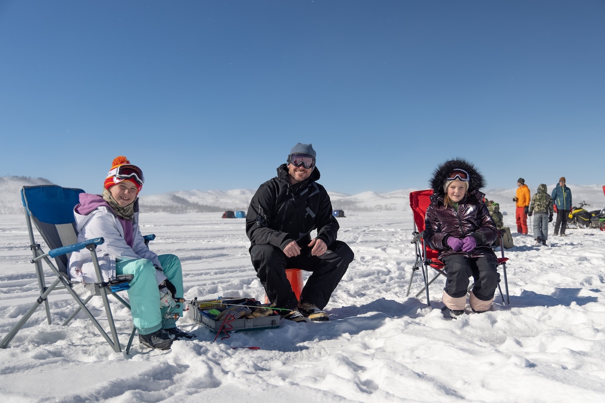 An adult and two children wearing winter coats sit on a frozen lake while ice fishing in Granby, Colorado.