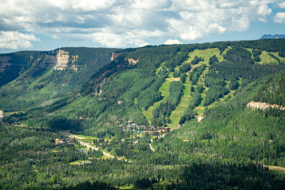 An elevated view of Purgatory Resort shows tree-lined ski runs, mountain cliffs and forests stretching across the San Juan Mountains in summer.