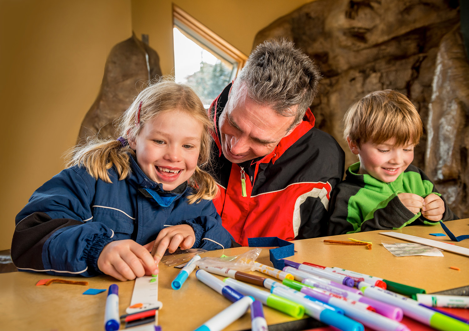 An adult in a red and black jacket sits between two children with blonde hair ad they complete a craft with markers and stickers in Snowmass, Colorado.