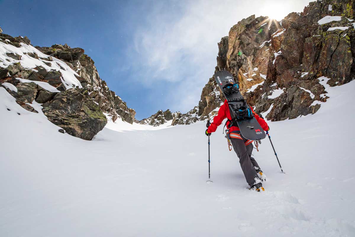 A backcountry adventurer treks uphill in the snow between two craggy rock walls in Colorado. They have a snowboard strapped onto their back.