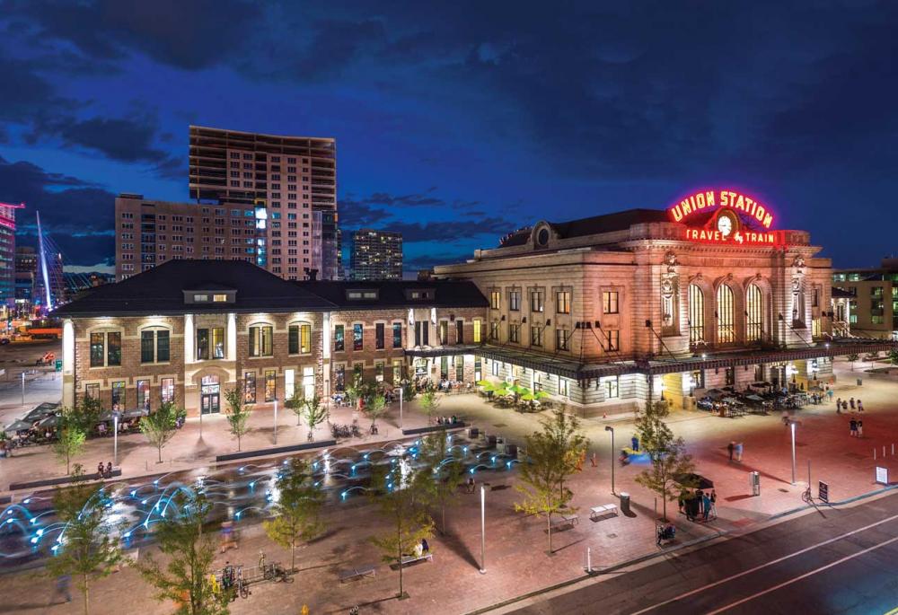Denver's Union Station at night. The historic building's red neon sign reads "Union Station" above a clock and below the clock it says "Travel By Train." There are small green trees surrounding a square with a water feature. There are people mingling around the station. And the blue night sky features some high rise buildings behind the building.
