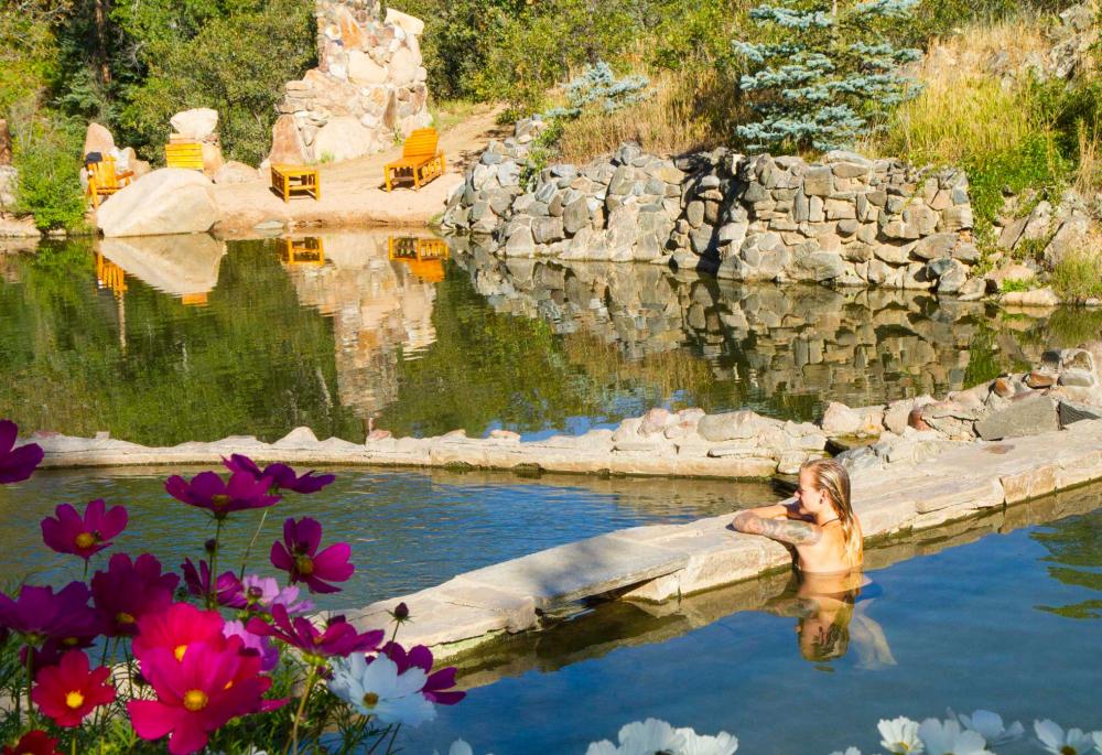 A woman sits in a hot springs looking out onto another body of water surrounded by a low rock wall. In the background stones lead to the waterfront with wooden chairs on a sandy beach.