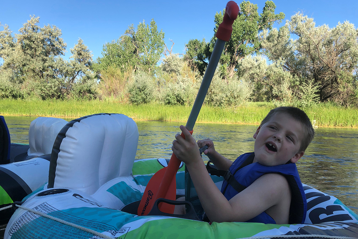 A child in a blue life jacket sits in a tube on a river with a green river bank with trees in the background.