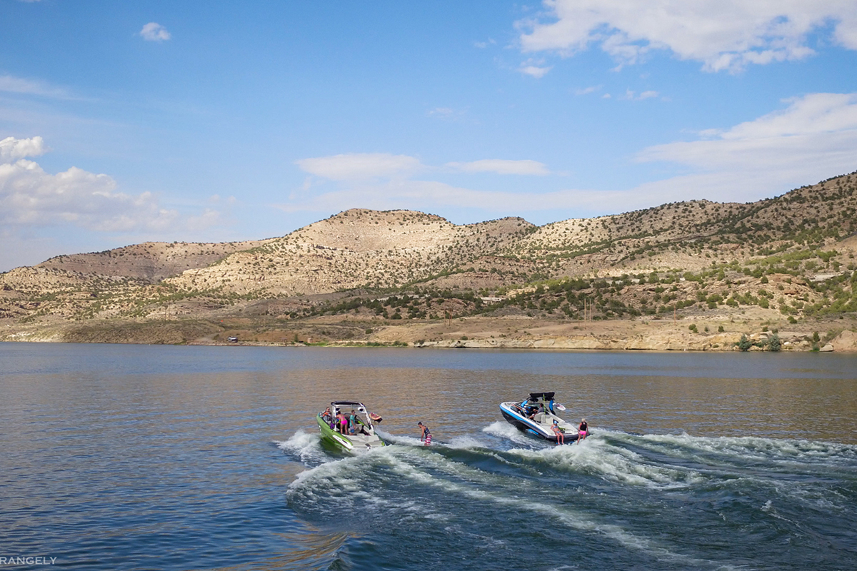 Two boats speed through the water at Kenney Reservoir with sand-colored hills in the background.