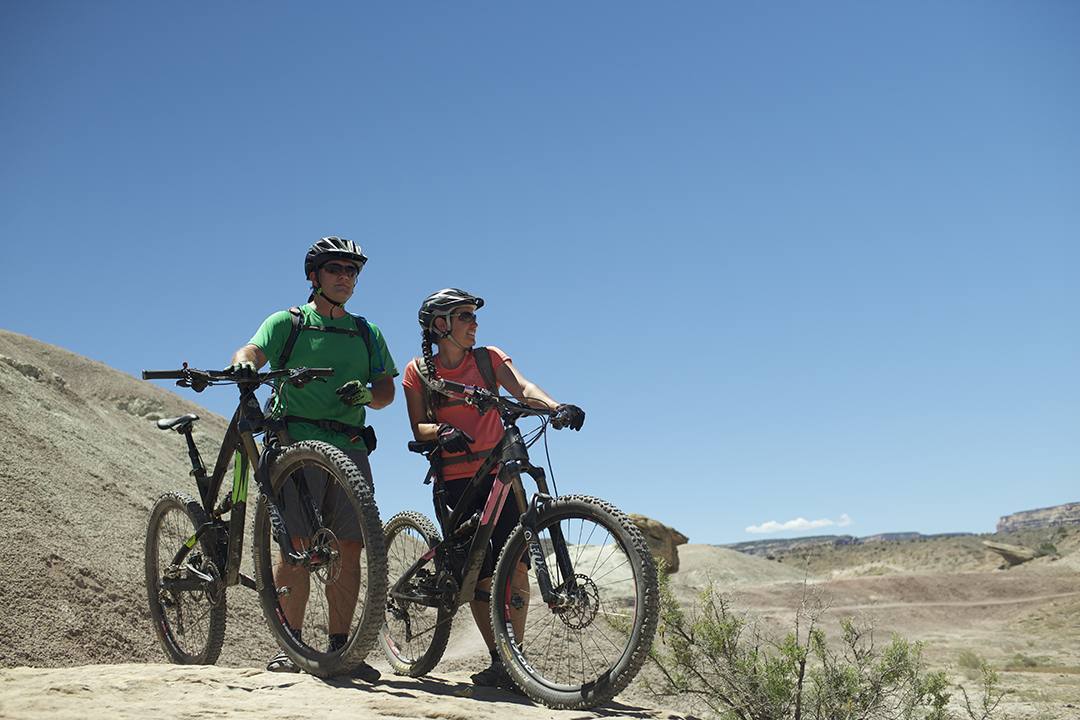 Two mountain bikers in helmets standing on a dirt landscape in Grand Junction, holding onto their bikes and looking out into the distance. The sky above them is a dusty blue.