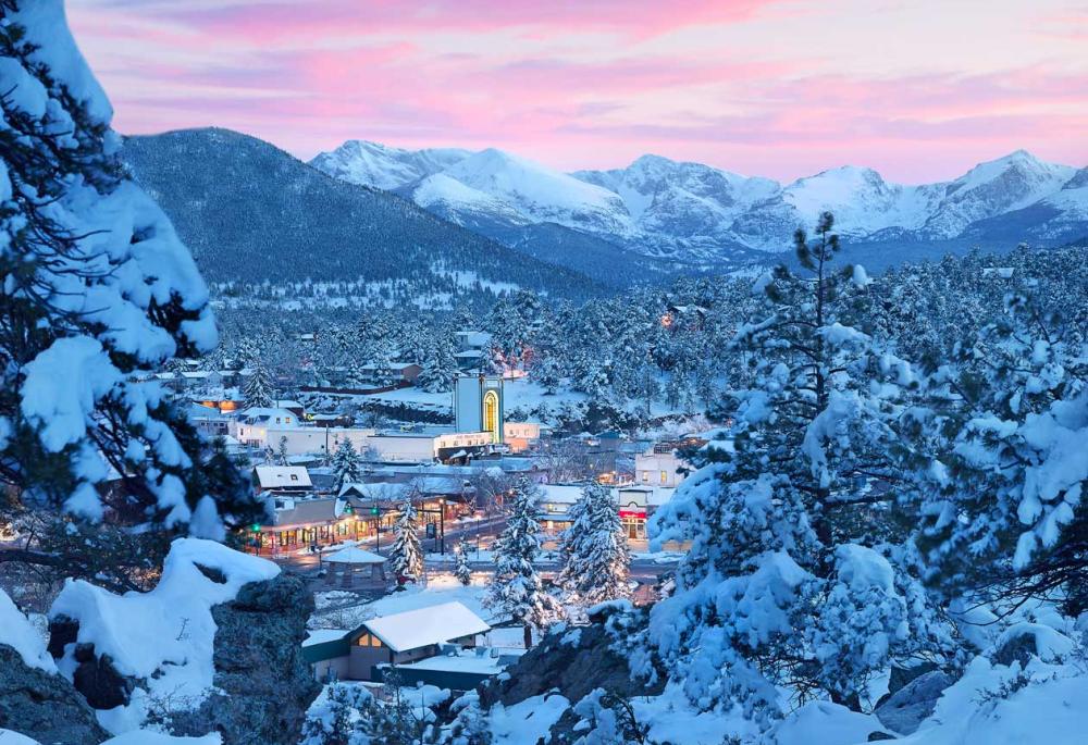 A snowy scene of Estes Park from a view point. The trees are coated in sugary white snow and the sky is a pink hue. The lights have come on in town.