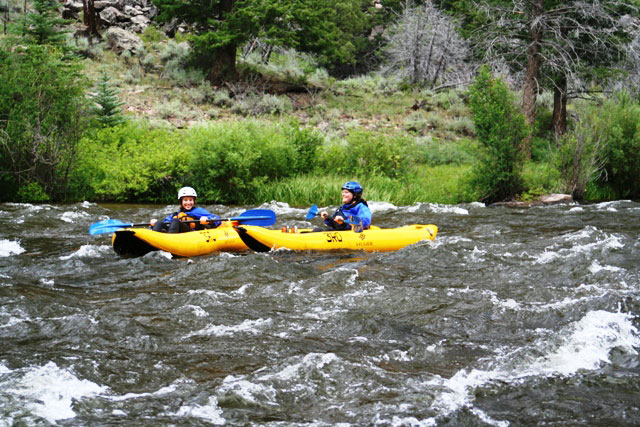 Two people in whitewater kayaks are paddling through white water rapids surrounded by greenery.