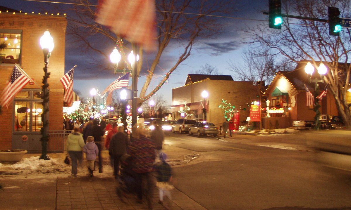 Olde Town Arvada bustling at night with many people walking the streets, shopping, dining and meeting with friends. The ground has snow and there are Christmas lights on buildings.