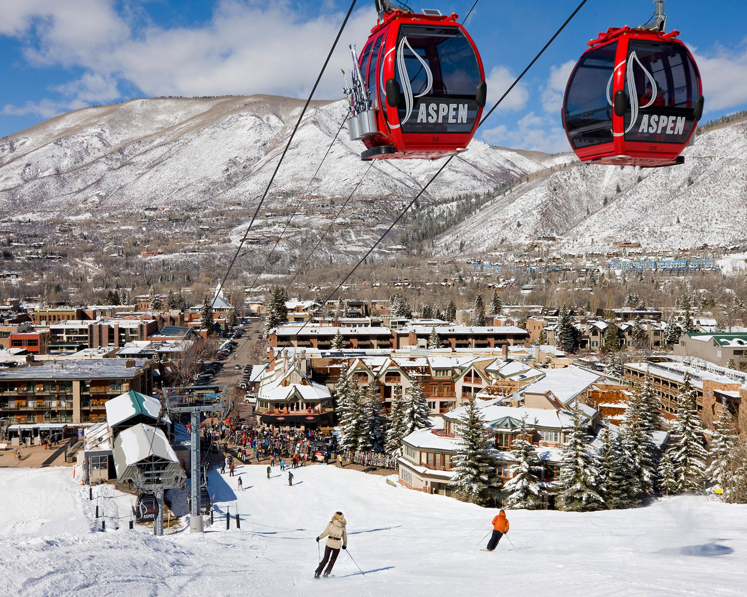 Two red and black Aspen Snowmass gondolas dangle over the ski area with hotels, mountains and a bright-blue sky in the background. Everything is covered in a layer of snow.