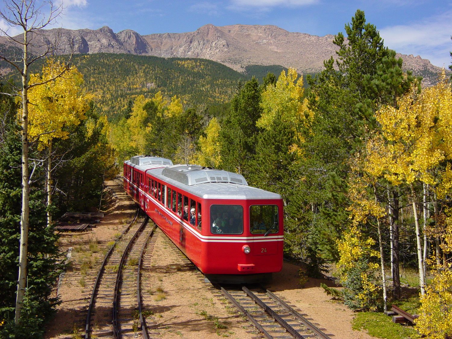 A small, modern red train with a silver roof and no smokestack chugs along a forest-lined track in Colorado. Aspens in the forest show off their golden hues. 