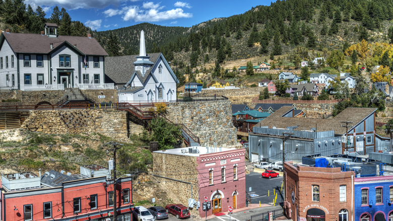 Historic, downtown Black Hawk on a bright, blue-sky summer’s day. Evergreen trees are in the background, covering mountains. Along a stone wall, a light blue historic church sits above old Western two story buildings. 