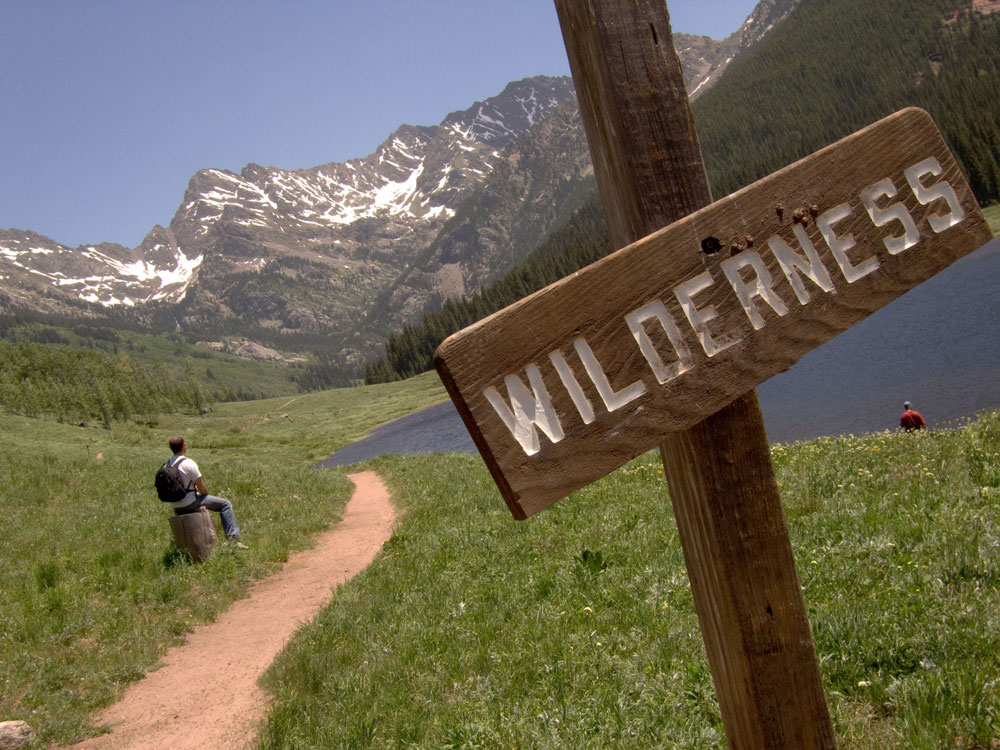 A sign that says "Wilderness" is posted next to a trail that runs through a green valley and next to a small blue lake. Snowcapped peaks rise in the distance