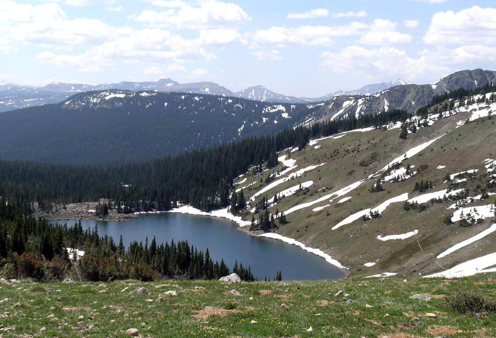 A navy-blue lake near Fort Collings sits calm and placid surrounded on three sides tall evergreen. On the last side is a steep, almost barren mountainside with streaks of spring snow.